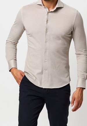 Homme portant une chemise beige à manches longues boutonnée avec col et un pantalon bleu marine foncé, main dans la poche, sur un fond clair uni.