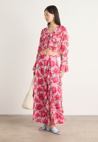 Set floreale rosa composto da un top corto con maniche lunghe a volant e una gonna abbinata, entrambi decorati con grandi fiori rossi e foglie verdi.