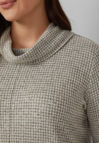 Frau in einem grauen, strukturierten Strickpullover mit weitem Rollkragen, vom Hals bis zur Mitte des Oberkörpers vor einem schlichten Hintergrund abgebildet.