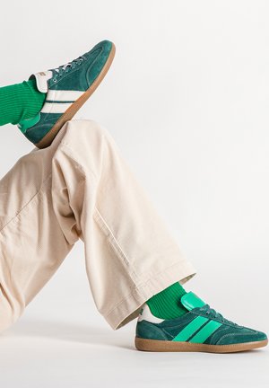 Personne portant un pantalon beige, des chaussettes vertes côtelées et des baskets vertes à bandes blanches, posant avec une jambe pliée et l'autre tendue.