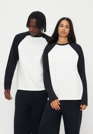 Dos personas que llevan camisetas raglán de manga larga en blanco y negro y pantalones negros están de pie frente a un fondo blanco liso.
