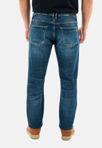 Jeans de denim azul oscuro con corte recto, con dos bolsillos traseros y una sutil textura descolorida. Llevados con zapatos de color marrón claro.
