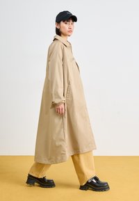 Casaco bege longo com um bolso frontal, combinado com calças largas cor areia e sapatos pretos de plataforma grossa. Modelo usando um boné preto. Fundo amarelo.