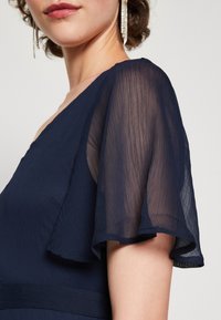 Mörkblå klänning med en flödande, genomskinlig flältsärm. Texturerad tyg med veckad design. Subtil detalj vid midjan förbättrar silhuetten.