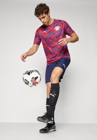Rød og blåmønstret Puma fotballdrakt, marineblå shorts, svarte sokker og svarte fotballsko, med en svart og hvit fotball som blir jonglert.