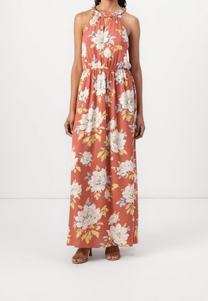 Frau trägt ärmelloses Maxikleid in rost-orange mit weiß-gelbem Blumenmuster und geflochtenem Neckholder, kombiniert mit braunen offenen Sandaletten.