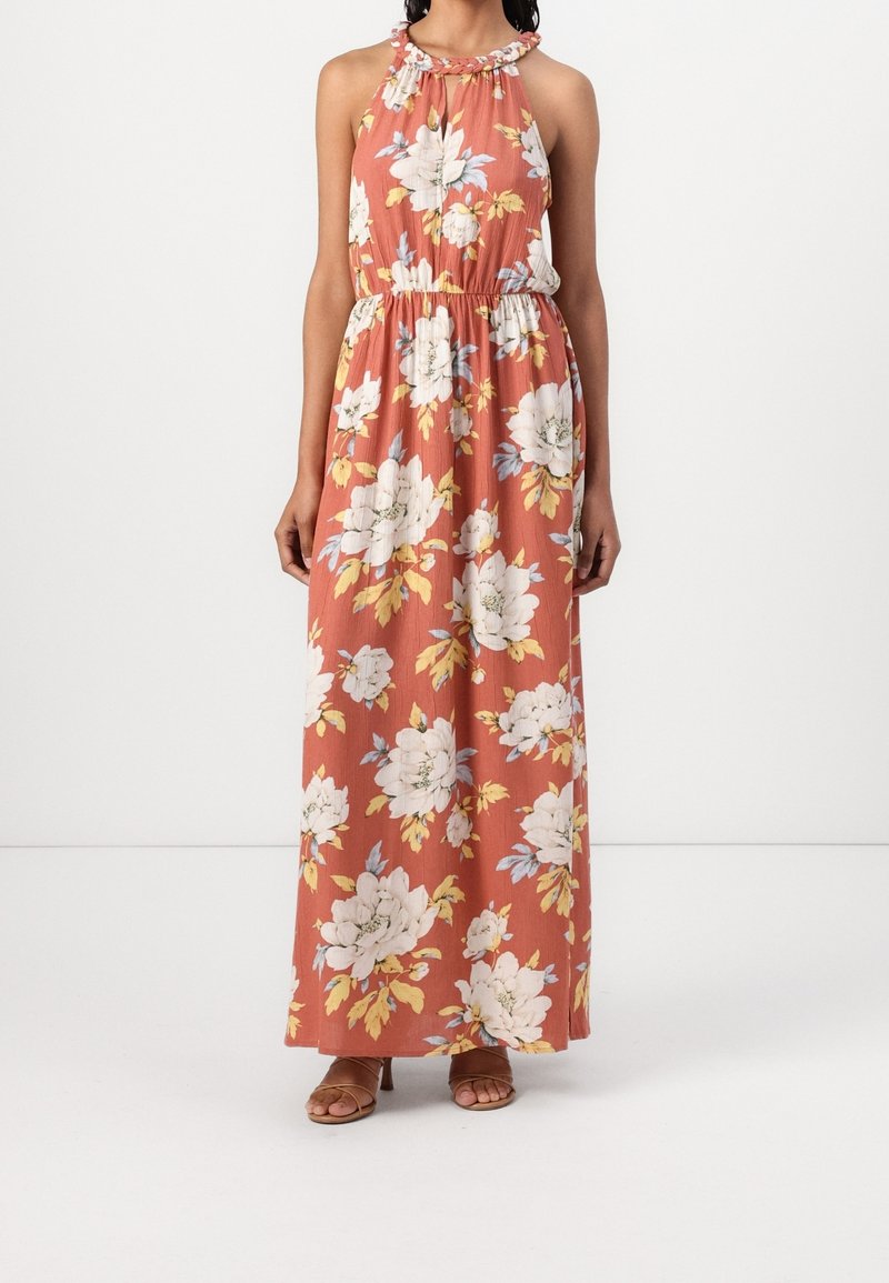 Femme portant une robe longue sans manches orange rouille avec imprimé floral blanc et jaune et col halter tressé, associée à des talons ouverts marron.