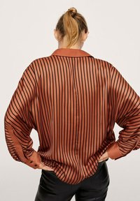 Frau mit blonden Pferdeschwanz, die eine locker sitzende, rostfarbene Bluse mit vertikalen schwarzen Streifen und schwarze Hose trägt, mit Händen in den Hüften, Rückansicht.