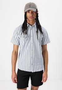 Chemise à manches courtes avec des rayures verticales bleues et blanches, devant à boutons, et une texture légère, accompagnée de shorts noirs et d'une casquette grise.
