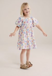 Abito floreale con maniche a sbuffo, caratterizzato da fiori multicolori su uno sfondo chiaro. Orlo a balze a più strati, abbinato a sandali beige.