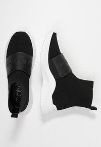 Černé pletené slip-on tenisky s bílou gumovou podrážkou. Vyznačují se texturovaným svrškem a širokým černým koženým pásem přes střední část nohy.