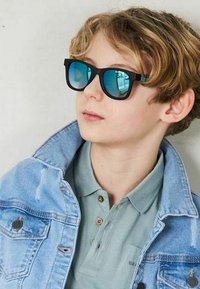 Svarta solglasögon med blå reflexlinser som bärs av ett barn med lockigt blont hår, ihop med en ljusgrön polo och en jeansjacka.