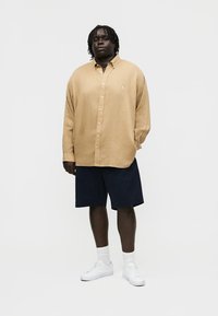 Vyras tamsios odos, su dredais, dėvintis šviesiai rudos spalvos ilgomis rankovėmis marškinius su sagutėmis, juodus šortus, baltas kojines ir baltus sportinius batelius, stovintis prieš baltą foną.