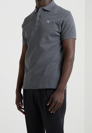 Dunkelhäutige Person trägt ein dunkelgraues Barbour-Poloshirt mit kurzen Ärmeln und schwarze Hosen und steht vor einem einfarbigen hellen Hintergrund.