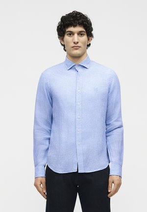 Νέος άνδρας με σγουρά μαλλιά που φοράει ανοιχτό μπλε πουκάμισο με κουμπιά και σκούρο παντελόνι, στέκεται μπροστά από ένα απλό λευκό φόντο.