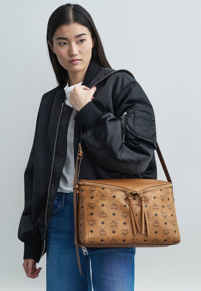 Junge Frau trägt eine schwarze Bomberjacke und blaue Jeans und hält eine große hellbraune Ledertasche mit gemusterten Logos über der Schulter.