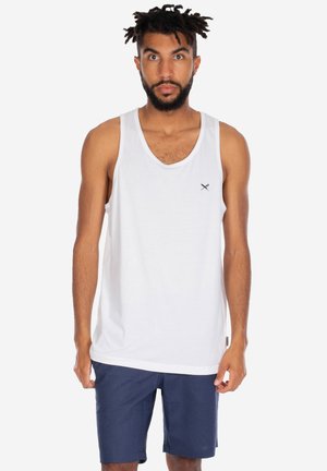 Uomo con la barba e dreadlocks corti che indossa una canottiera bianca e pantaloncini blu navy, in piedi su uno sfondo bianco semplice.