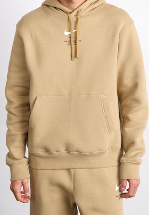 Beżowa bluza z kapturem Nike i dopasowane spodnie dresowe noszone przez osobę, z małymi białymi logo Nike Swoosh oraz przednią kieszenią na bluzie.