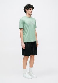 Zöld pamut póló "Emporio Armani" logóval, fekete rövidnadrággal, amelyen ismétlődő márkajelzés található. A modell fehér tornacipőt és zoknit visel.