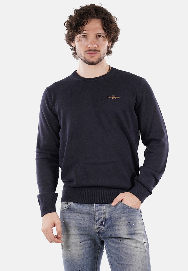 Maglione di cotone navy con collo rotondo, polsini e orlo a coste. Presenta un logo ricamato sul petto. Abbinato a jeans blu sbiaditi.