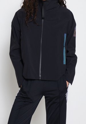 Person trägt schwarze Jacke mit Reißverschluss und hohem Kragen sowie schwarze Hose mit weißen Streifen, Hände in den Taschen, vor weißem Hintergrund.