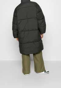 Donkergrode pufferjas met een lange, oversized pasvorm, voorzien van doorstikte secties. In combinatie met olijfgroene wijde broekspijpen en witte sneakers.