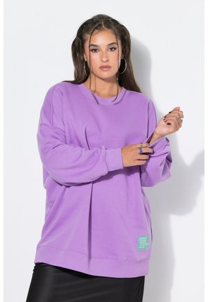 Femme aux longs cheveux bruns et créoles portant un sweatshirt lavande oversize et une jupe noire, debout sur un fond blanc uni.