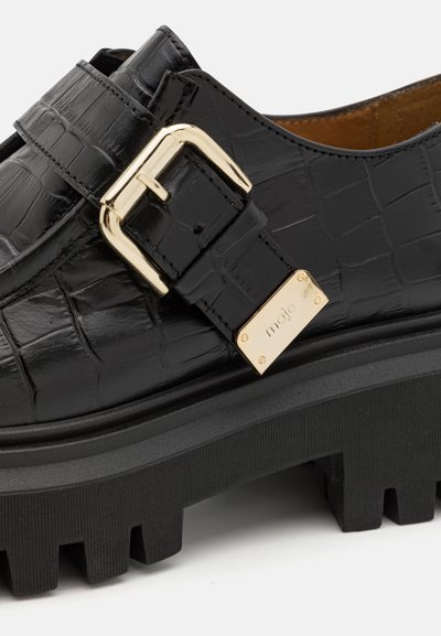 Scarpa nera in ecopelle con texture a crocodrillo, accentuata da una fibbia dorata e suola nera spessa e rigata. Dettaglio hardware notevole visibile.