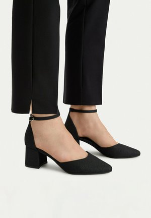 Pies con zapatos negros de tacón grueso con tiras en el tobillo combinados con pantalones negros recortados con aberturas en la parte trasera.