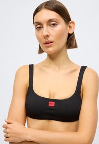 Črn rebrast bikini zgornji del z širokimi naramnicami, kvadratnim izrezom in rdečo značko z logotipom na sredini spredaj. Mehka tekstura in oprijeta zasnova.