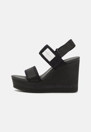 Czarna sandałka na koturnie z dwoma szerokimi paskami z tkaniny, paskiem na kostkę na rzep z białą metką Calvin Klein Jeans i podeszwą na platformie.