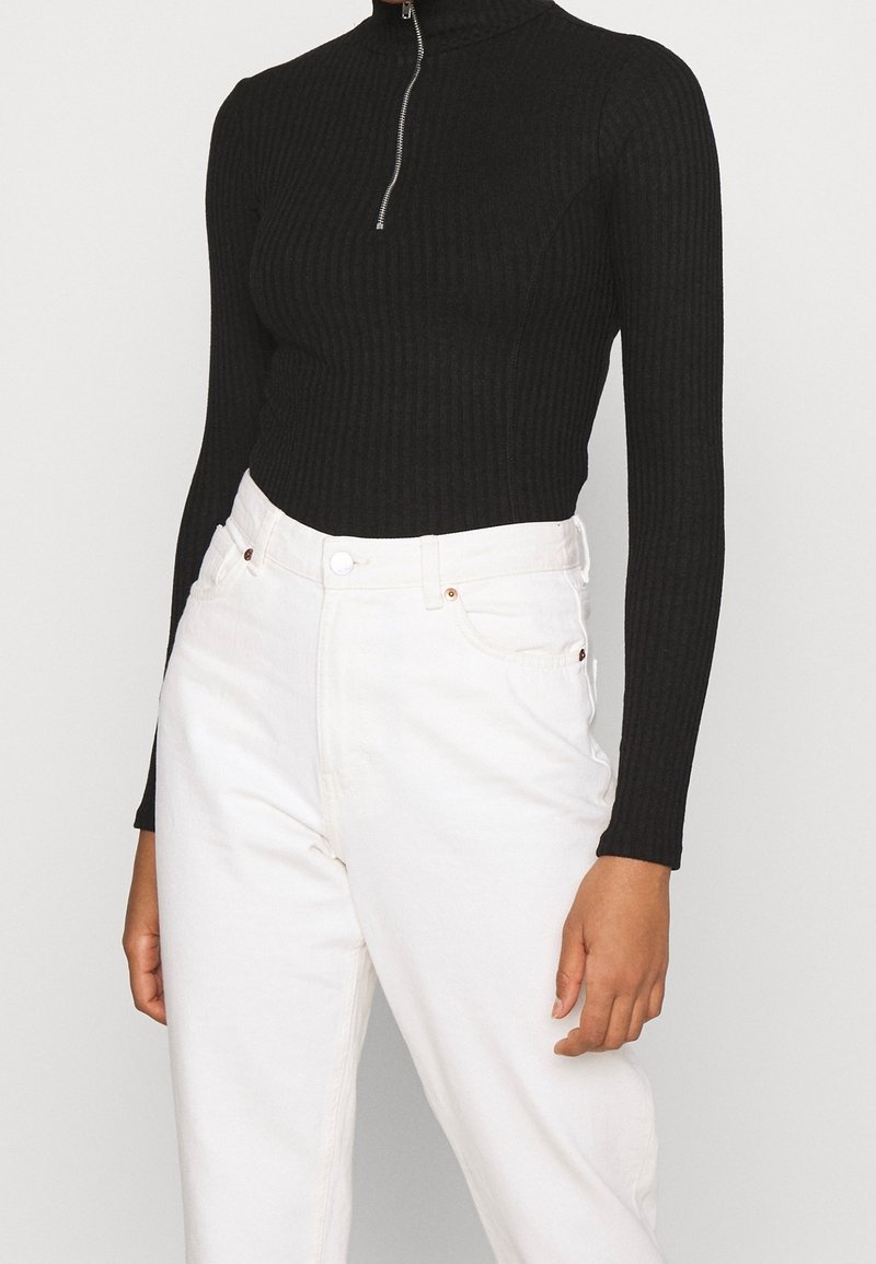 Černý žebrovaný top s dlouhými rukávy a vysokým límcem se stříbrným zipem, kombinovaný s bílými džínami s vysokým pasem.