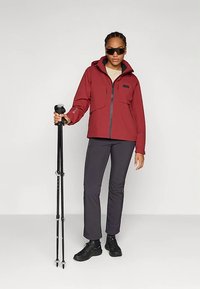 Rode waterdichte jas met een ritssluiting aan de voorkant, twee borstzakken, gedragen met een grijze broek en zwarte schoenen. De persoon houdt een wandelstok vast.