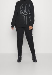 Pull noir avec motif de visage abstrait blanc, col et poignets côtelés, associé à un pantalon noir et des bottines. Texture lisse, coupe oversize.