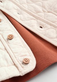 Quilted beige fabric with terracotta panel, featuring round wooden buttons and ribbed texture. Close-up view highlighting stitching details.