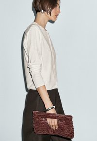 Donna con i capelli corti, veste un cardigan chiaro e pantaloni scuri, tiene in mano una borsa a tracolla intrecciata di colore marrone ed indossa un bracciale d'argento, posata su uno sfondo chiaro.