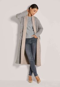 Frau, die einen langen grauen und beigen Mantel über einem grauen Pullover und dunklen Skinny Jeans trägt, steht mit einer Hand am Nacken und der anderen in der Tasche.