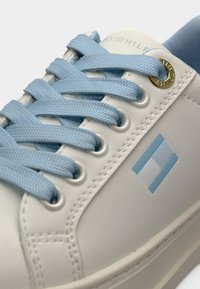 Λευκό συνθετικό αθλητικό παπούτσι με υφή στην επιφάνεια, ανοιχτό μπλε κορδόνια, χρυσής απόχρωσης μεταλλική τρύπα κορδονιών και μπλε γεωμετρικό λογότυπο στο πλάι.