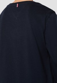 Granatowa koszulka z długim rękawem, wykonana z miękkiej bawełny, z małym trójkolorowym akcentem z tyłu przy dekolcie oraz klasycznym okrągłym dekoltem.