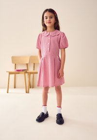 Vestito a quadretti rossi e bianchi con colletto alla Peter Pan, maniche corte a sbuffo e bottoni frontali. Abbinato a scarpe nere e calzini bianchi.