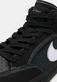 Czarny wysokoprofilowy sneaker z grubymi czarnymi sznurowadłami, białym przeszyciem, białym logo Nike i napisem „Leo Baker” na języku.