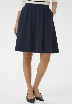 Kvinde iført en marineblå nederdel til knæet med knapper ved taljebåndet og sorte spidse hæle.