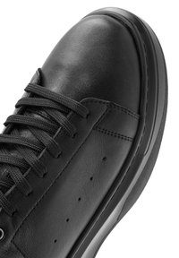 Zwarte leren sneaker met zwarte veters, zichtbare stiksels en kleine ventilatiegaten aan de zijkant, weergegeven in een close-up en schuin uitzicht.