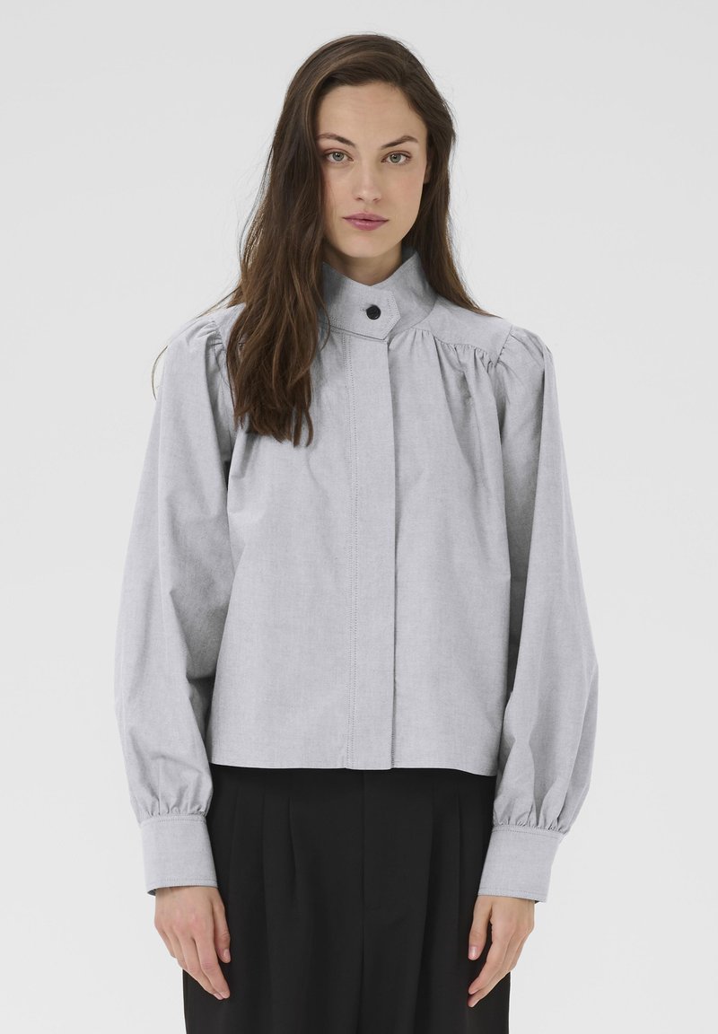 Kvinde med langt brunt hår iført en lys grå knapbluse med puffede ærmer og sorte vidde bukser, stående mod en ensfarvet baggrund.