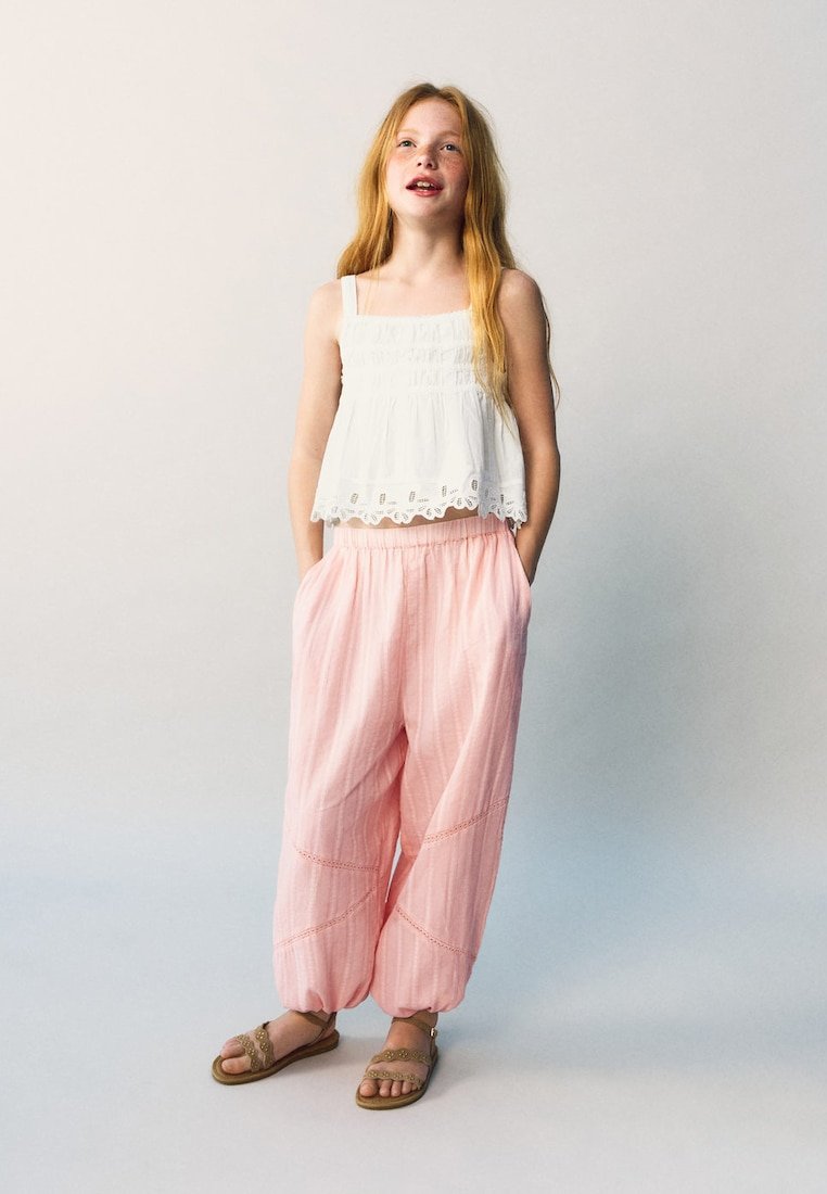 Jeune fille aux longs cheveux roux portant un haut blanc sans manches, un pantalon rose ample et des sandales marron, debout les mains dans les poches.