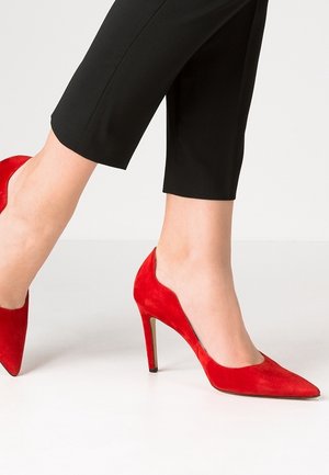 Rode suede hoge hakken met een spitse neus, slanke stiletto hak en een slanke silhoutte; gecombineerd met zwarte cropped pants.