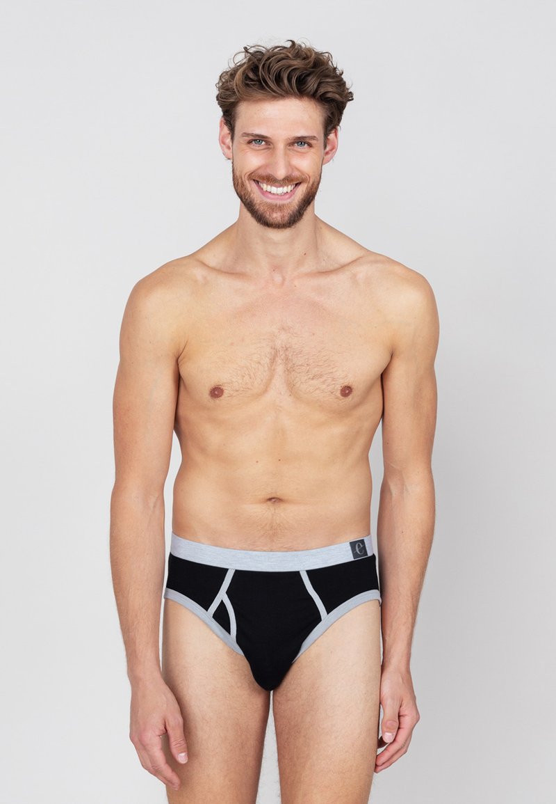 Zwarte katoenen slips met grijze elastische tailleband en accenten. Heeft een strakke pasvorm en een minimalistisch ontwerp. Model staat met een neutrale uitdrukking.