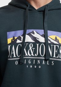 Dark green hoodie with a front graphic featuring mountains, trees, and sun in purple and yellow, branded "JACK & JONES ORIGINALS" in white text.