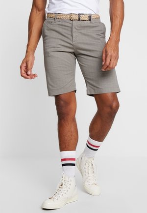 Homme portant un short beige texturé avec une ceinture tressée, des baskets blanches montantes et des chaussettes blanches à rayures rouges et noires.