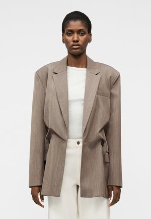 Osoba nosi nadrozmerné taupe sako cez biely vrch a krémové nohavice s vysokým pásom, stojaca pred jednoduchým svetlým pozadím.